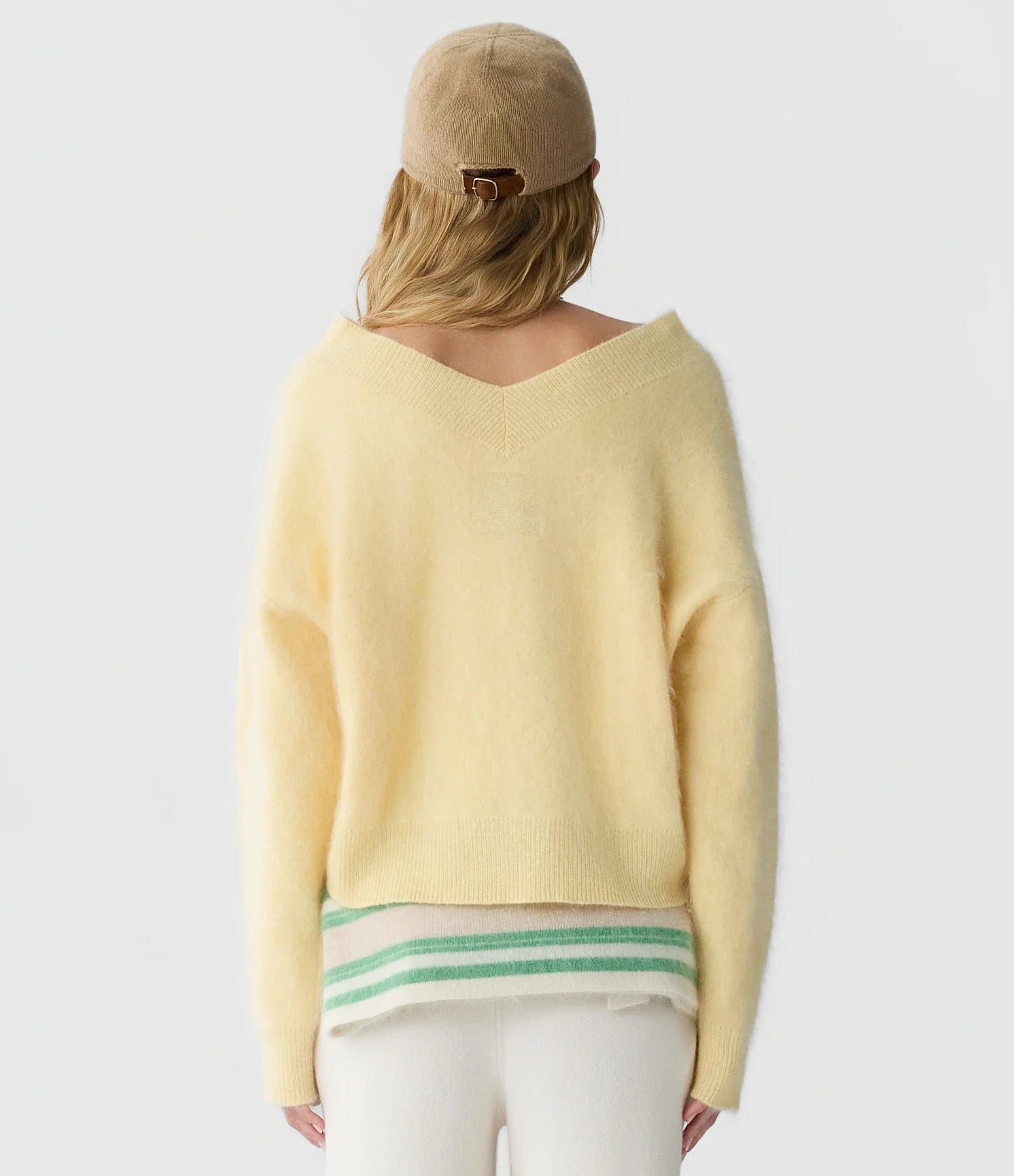 A person facing away wears the Grizzly Double V Sweater by Guest in Residence—a lightweight cashmere pullover in a subtle yellow shade with a double V neckline—styled with a beige cap, white pants, and a green-and-white striped layer visible underneath. The background is plain white.