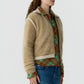 Woman wears the Grizzly Frankie Jacket by Guest in Residence over a green and brown argyle shirt with blue jeans, standing against a plain light background.