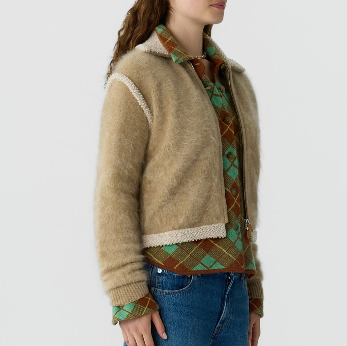 Woman wears the Grizzly Frankie Jacket by Guest in Residence over a green and brown argyle shirt with blue jeans, standing against a plain light background.