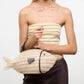 A person in a strapless striped top and dark pants holds the Staud Cleo Fish Bag, featuring a leather-wrapped handle.