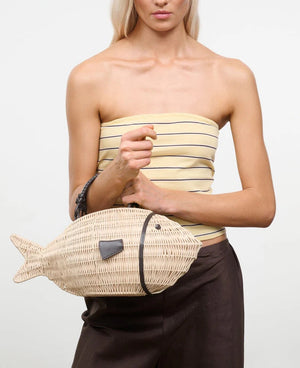 A person in a strapless striped top and dark pants holds the Staud Cleo Fish Bag, featuring a leather-wrapped handle.