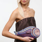 A woman with long blonde hair in a strapless brown dress holds the Staud Pesce Clutch, a large, beaded fish-shaped bag with a zip closure.
