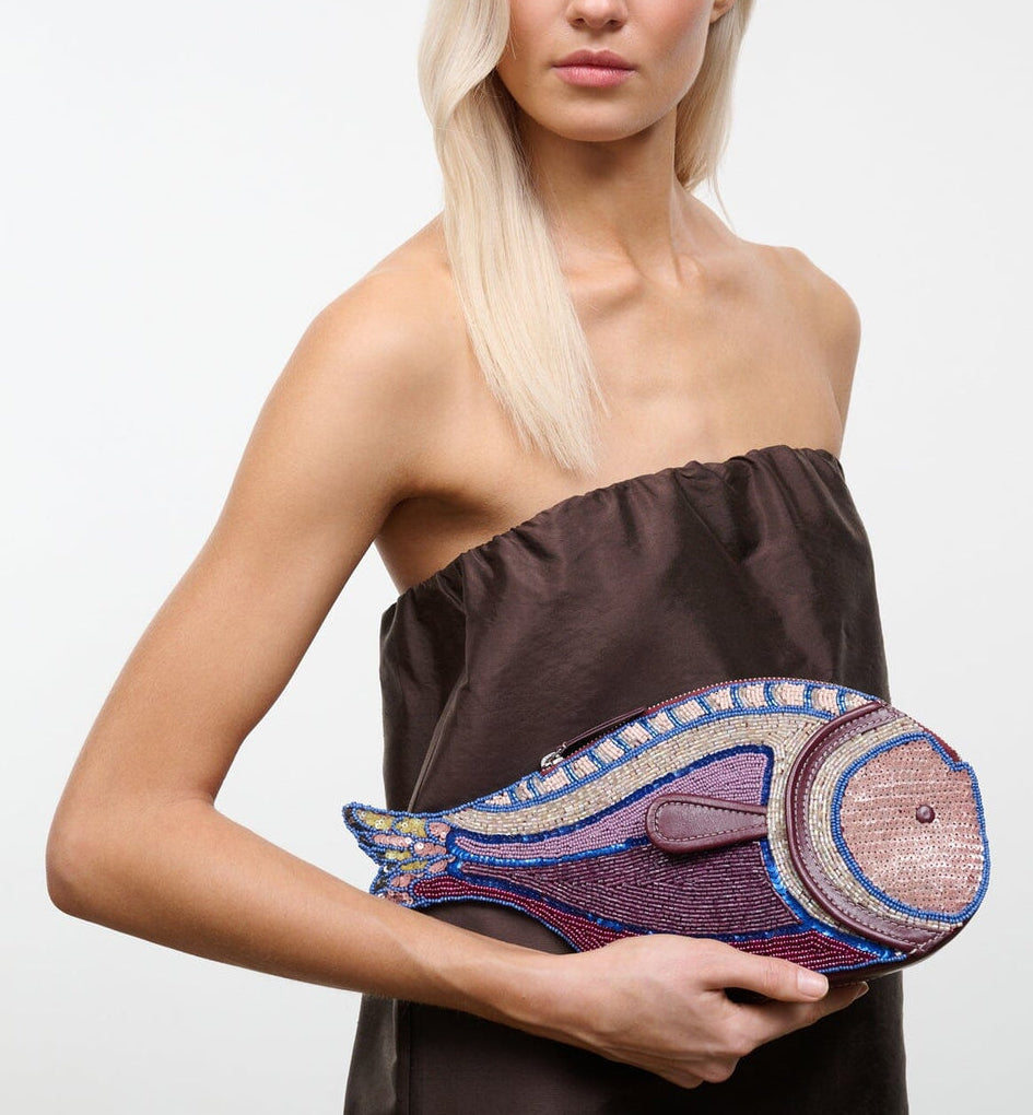A woman with long blonde hair in a strapless brown dress holds the Staud Pesce Clutch, a large, beaded fish-shaped bag with a zip closure.
