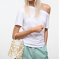 A woman in a white off-shoulder t-shirt and light green pants holds the Staud Lila Mini Bag, a gold sequin evening accessory.