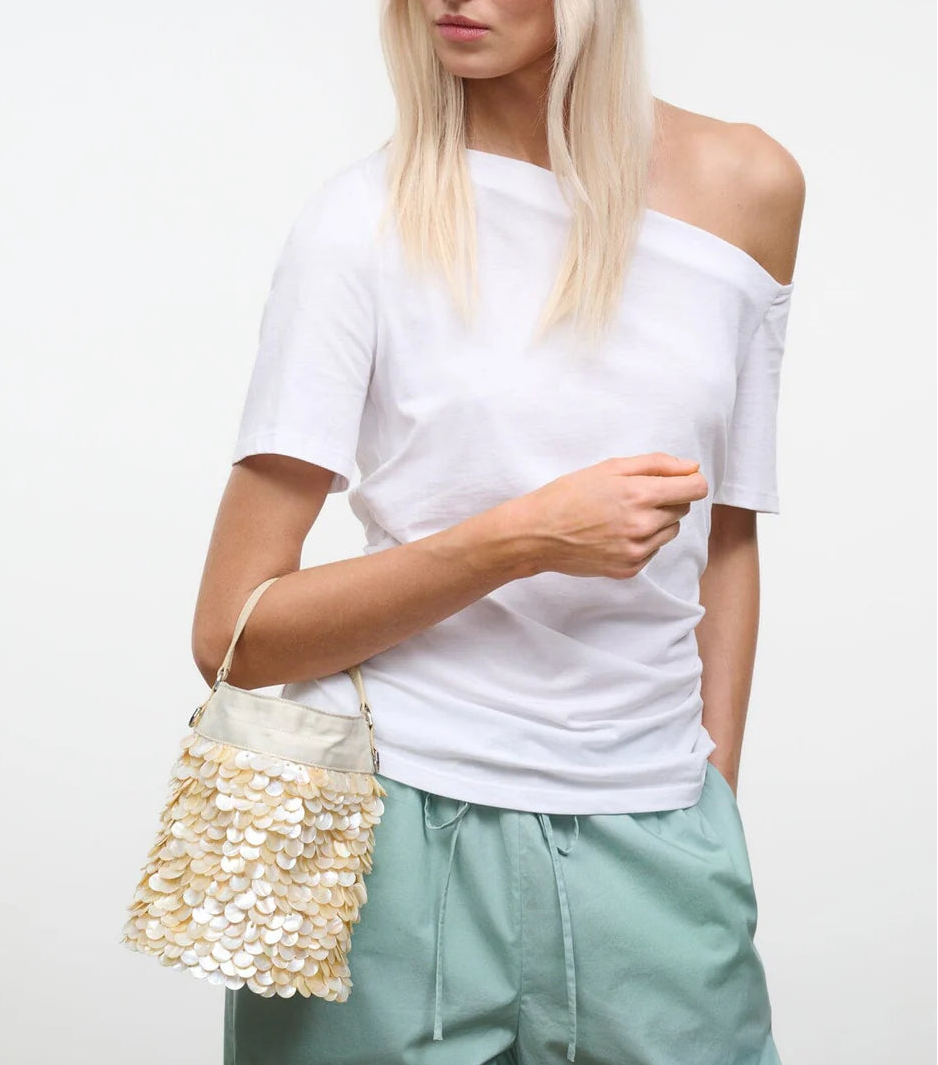 A woman in a white off-shoulder t-shirt and light green pants holds the Staud Lila Mini Bag, a gold sequin evening accessory.