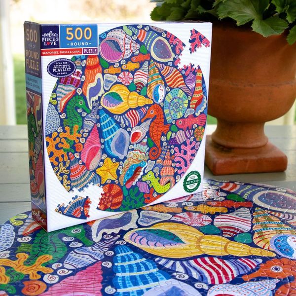 The Eeboo Seahorses, Shells & Coral 500 Round Puzzle by Monika Forsberg features colorful seashells, coral, and marine wonders, shown on a table beside a potted plant.