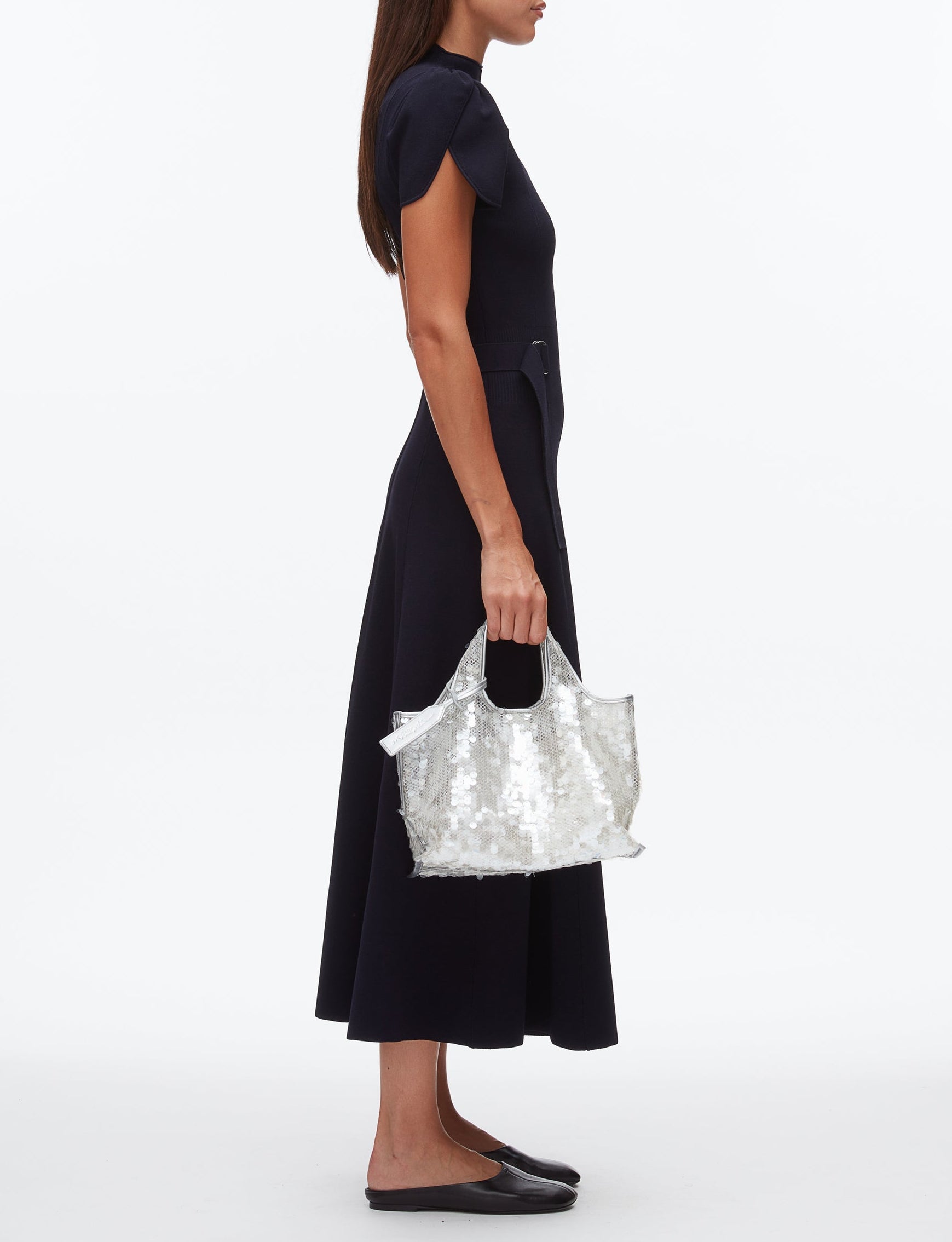 A woman in a black short-sleeve midi dress and black flats holds the 3.1 Phillip Lim Mesh Sequin Mini Market Tote, standing in profile against a plain white background.