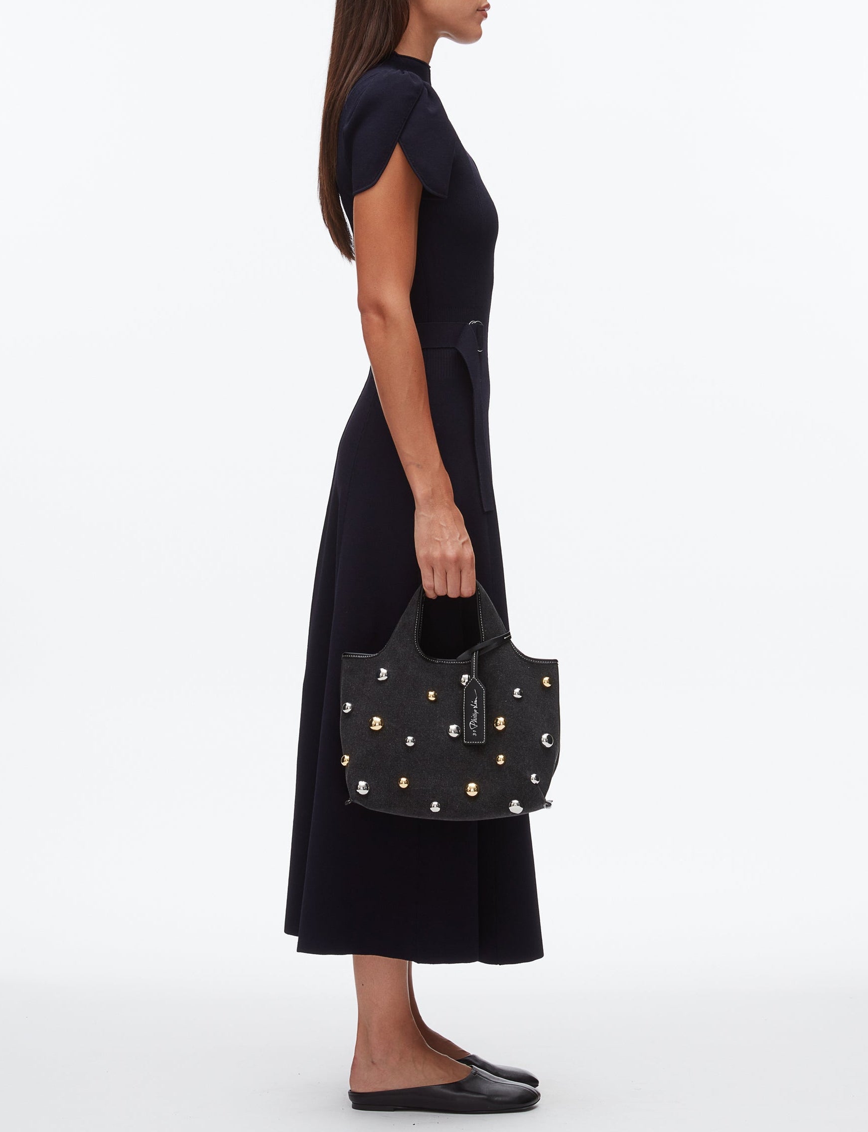 A woman in a black midi dress and flats stands in profile, holding the 3.1 Phillip Lim Washed Denim Mini Market Tote.