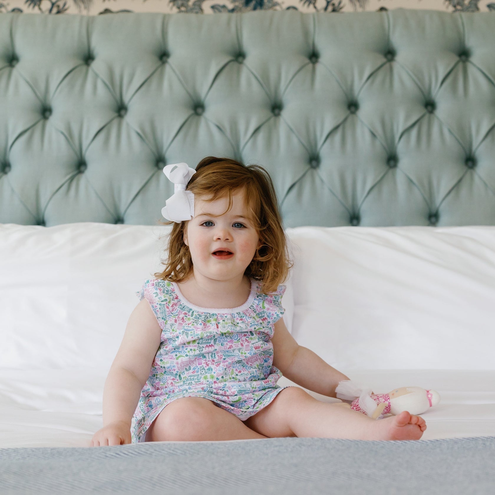 A young child, dressed in a Joy Street Girls' Emilia Dress with a lovely floral pattern inspired by sunny days in Palm Beach, Florida, sits on a bed with a tufted headboard. Made of soft Pima cotton, the dress adds to her comfort as she holds a doll and smiles. Her white bow adds an extra touch of charm to the scene.