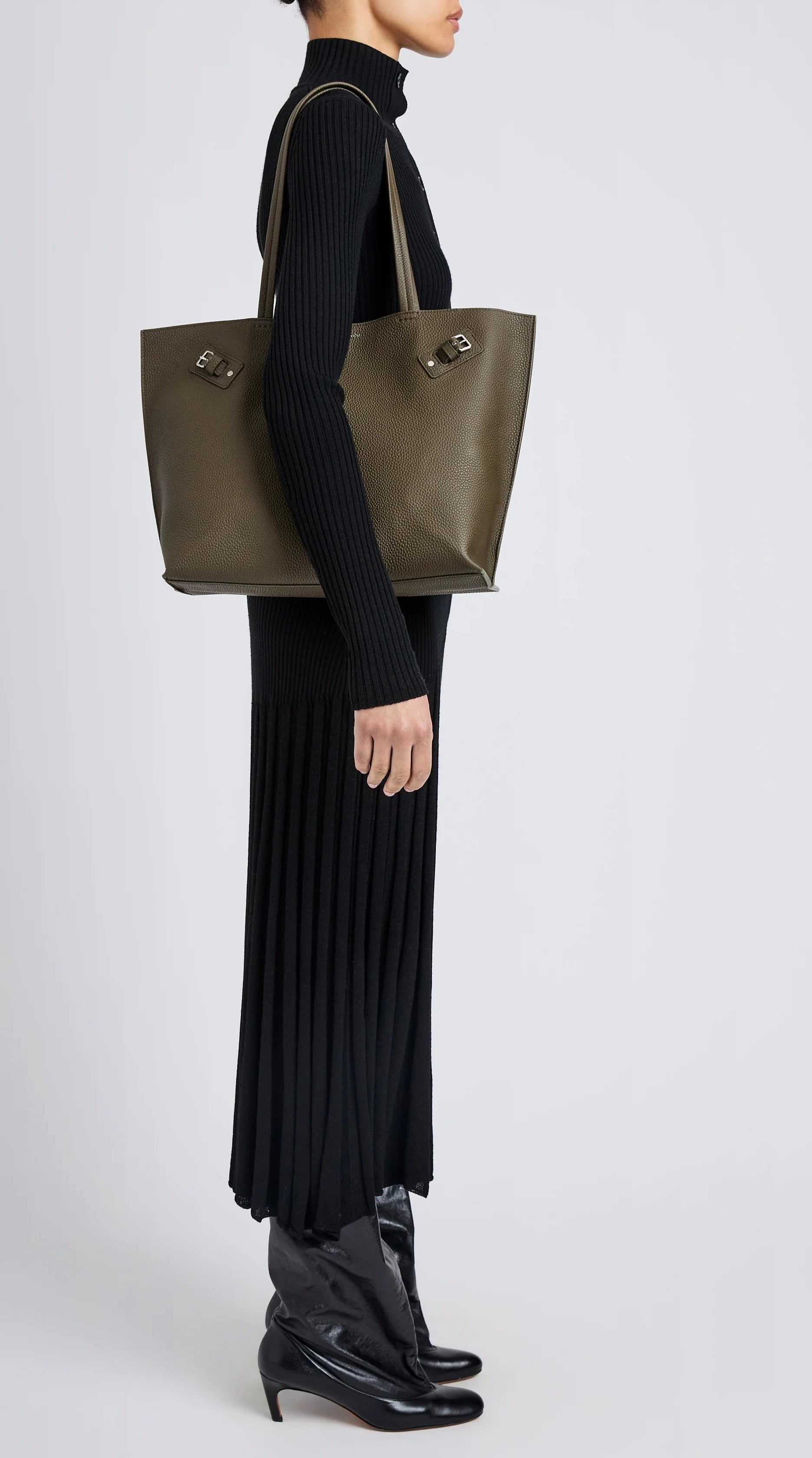 A person in a black ribbed turtleneck dress and black boots stands in profile, carrying the Proenza Schouler Days Pebbled Calf Skin Tote on their shoulder.