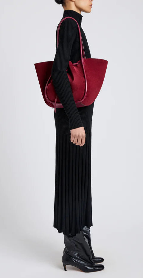 A person in a black long-sleeve pleated dress and black boots stands sideways, carrying a Proenza Schouler Large Ruched Soft Suede Tote with a gold-tone logo on their shoulder against a plain background.