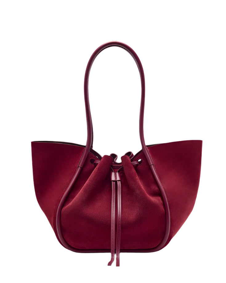 The Proenza Schouler Large Ruched Soft Suede Tote is a spacious, deep red bag with two rounded handles, a gold-tone logo, and a softly structured silhouette.