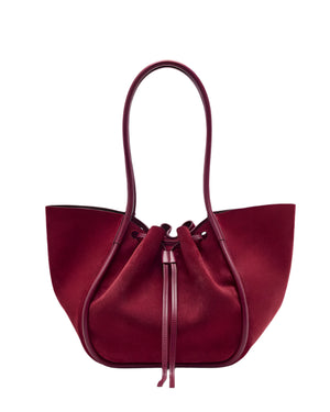 The Proenza Schouler Large Ruched Soft Suede Tote is a spacious, deep red bag with two rounded handles, a gold-tone logo, and a softly structured silhouette.