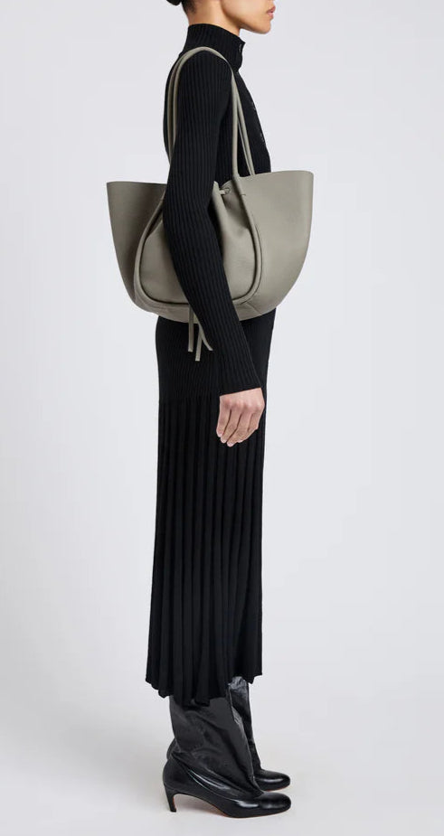 A person in a long black ribbed dress and black boots stands in profile, carrying the Proenza Schouler Large Ruched Soft Pebbled Calf Tote on their shoulder, with a leather zip pouch peeking out for added style.