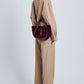A person in a loose beige pantsuit stands sideways, carrying the Proenza Schouler Small Ruched Soft Suede Tote in dark burgundy with a signature drawstring design.