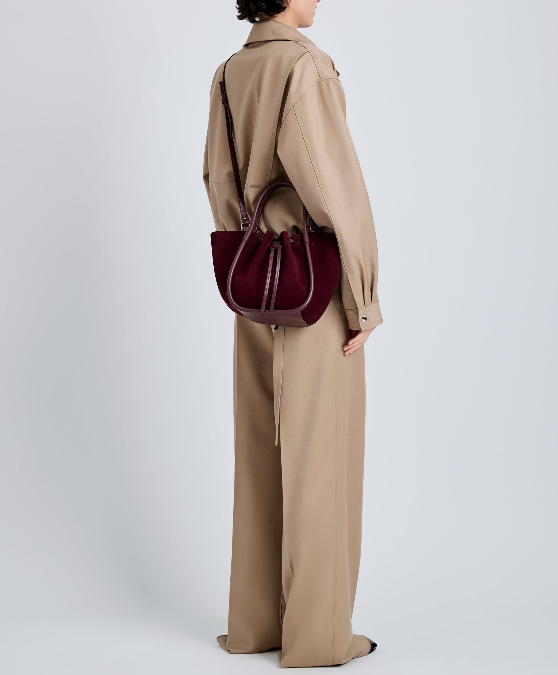 A person in a loose beige pantsuit stands sideways, carrying the Proenza Schouler Small Ruched Soft Suede Tote in dark burgundy with a signature drawstring design.