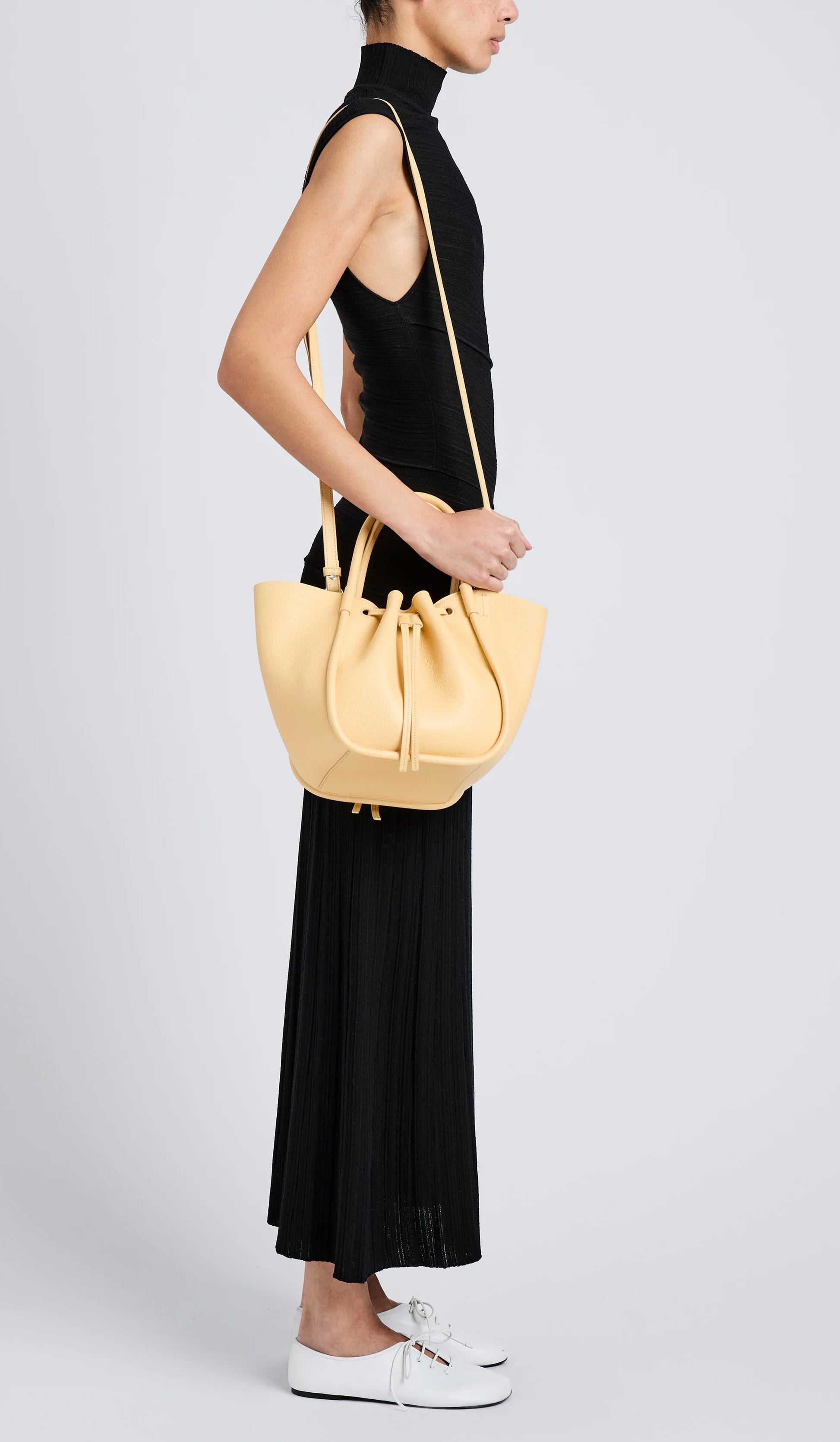 A woman in a sleeveless black dress and white slip-on shoes, shown in profile, carries a light yellow Proenza Schouler Small Ruched Soft Pebbled Calf Tote with a sculptural silhouette.