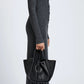 A person in a long, ribbed grey dress stands in profile, holding the Proenza Schouler Small Ruched Soft Pebbled Calf Tote and wearing black boots against a plain background.