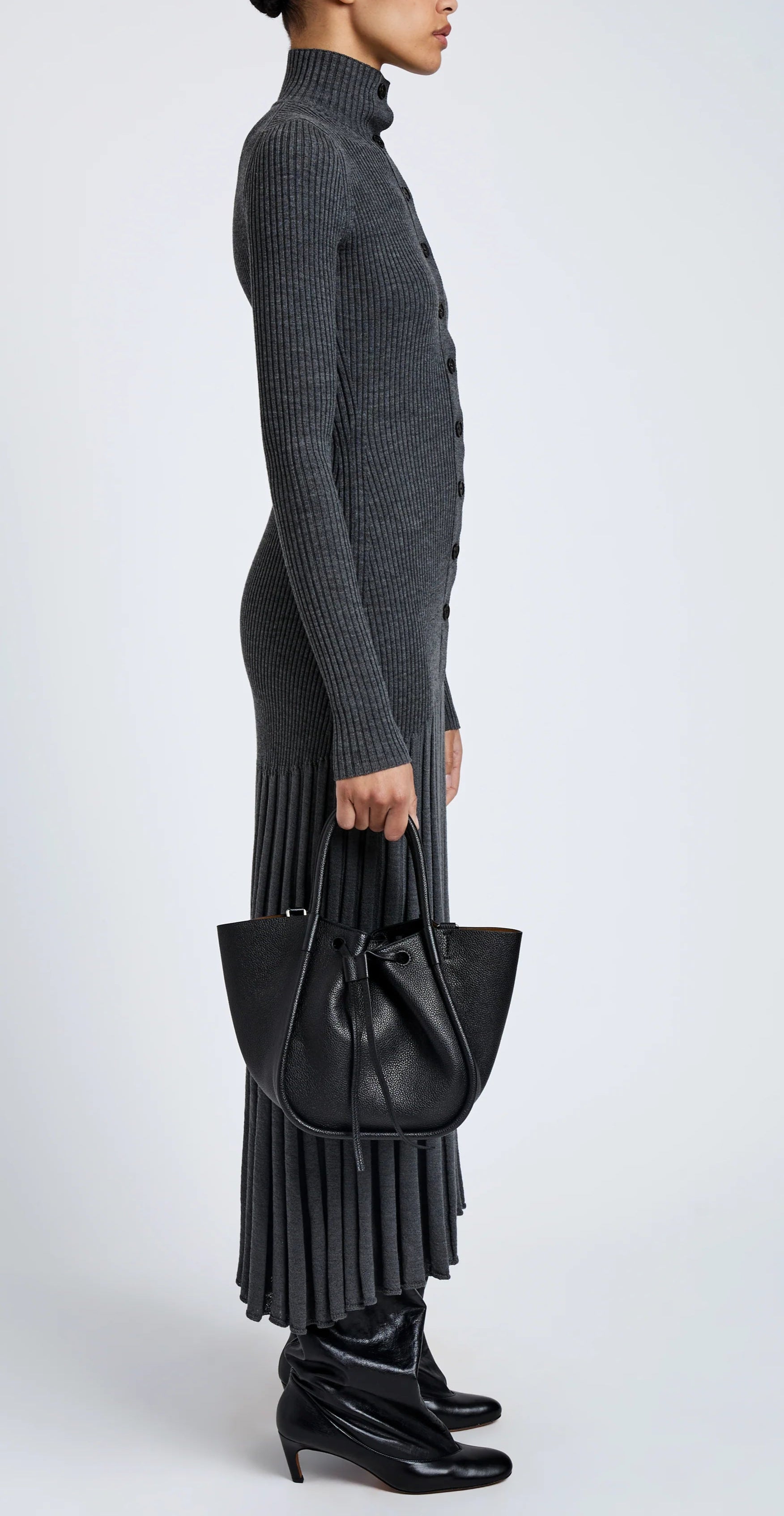 A person in a long, ribbed grey dress stands in profile, holding the Proenza Schouler Small Ruched Soft Pebbled Calf Tote and wearing black boots against a plain background.