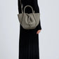 A person in a long black ribbed dress and black boots carries the Proenza Schouler Small Ruched Soft Pebbled Calf Tote, its sculptural silhouette with top handles and shoulder strap standing out against a plain light background.