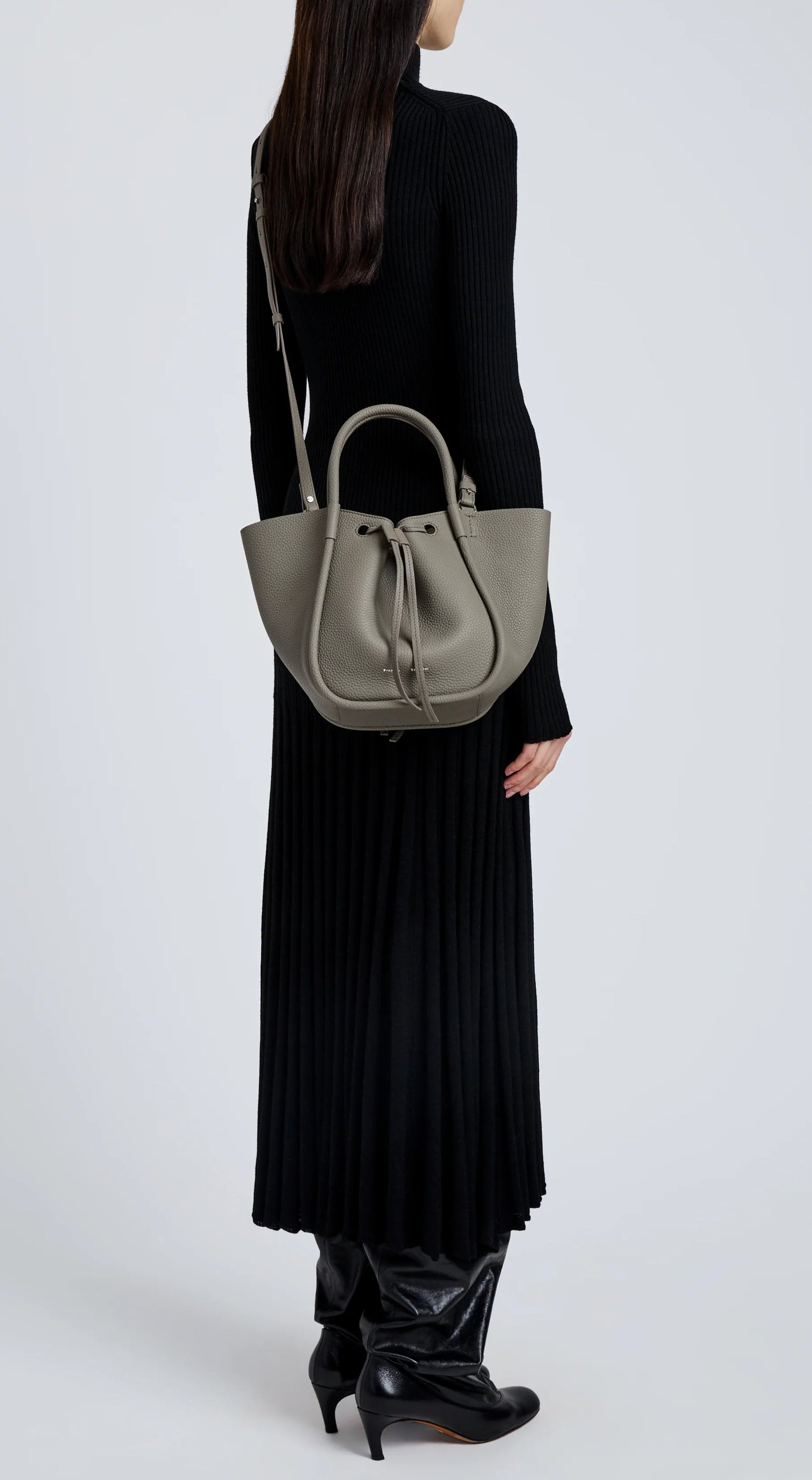 A person in a long black ribbed dress and black boots carries the Proenza Schouler Small Ruched Soft Pebbled Calf Tote, its sculptural silhouette with top handles and shoulder strap standing out against a plain light background.