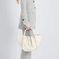 Person in a light gray pantsuit and silver heels holds the Proenza Schouler Small Ruched Soft Pebbled Calf Tote, featuring knotted details and a sculptural silhouette, against a plain light background.