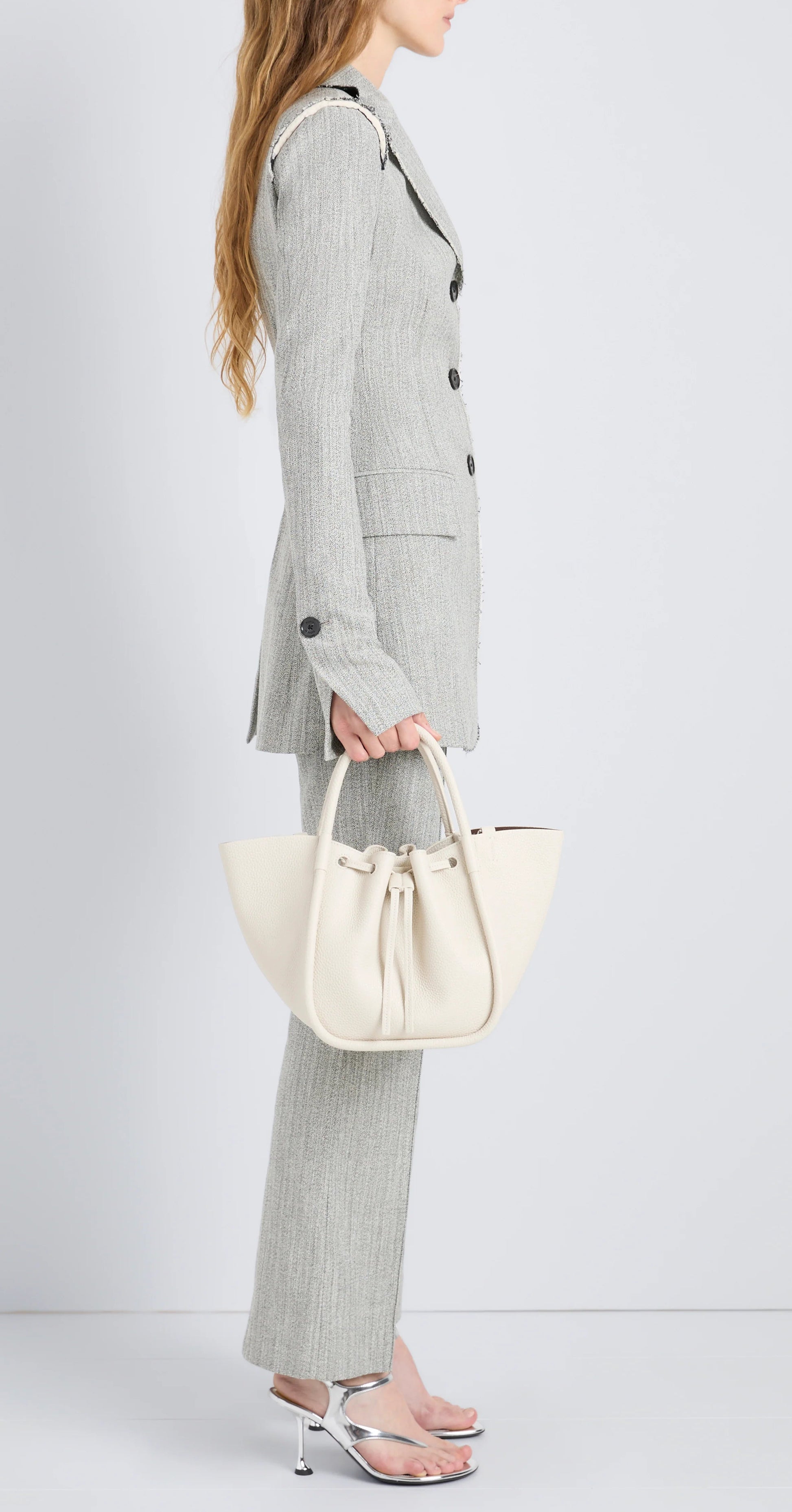 Person in a light gray pantsuit and silver heels holds the Proenza Schouler Small Ruched Soft Pebbled Calf Tote, featuring knotted details and a sculptural silhouette, against a plain light background.