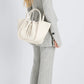 A person with long wavy hair in a grey pantsuit and heeled sandals stands sideways, carrying the Proenza Schouler Small Ruched Soft Pebbled Calf Tote with an open top and sculptural silhouette on their arm.