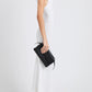 A person in a sleeveless white dress and white sneakers holds the Proenza Schouler Tate Clutch, a compact hand-held bag with a detachable crossbody strap, standing against a plain white background.