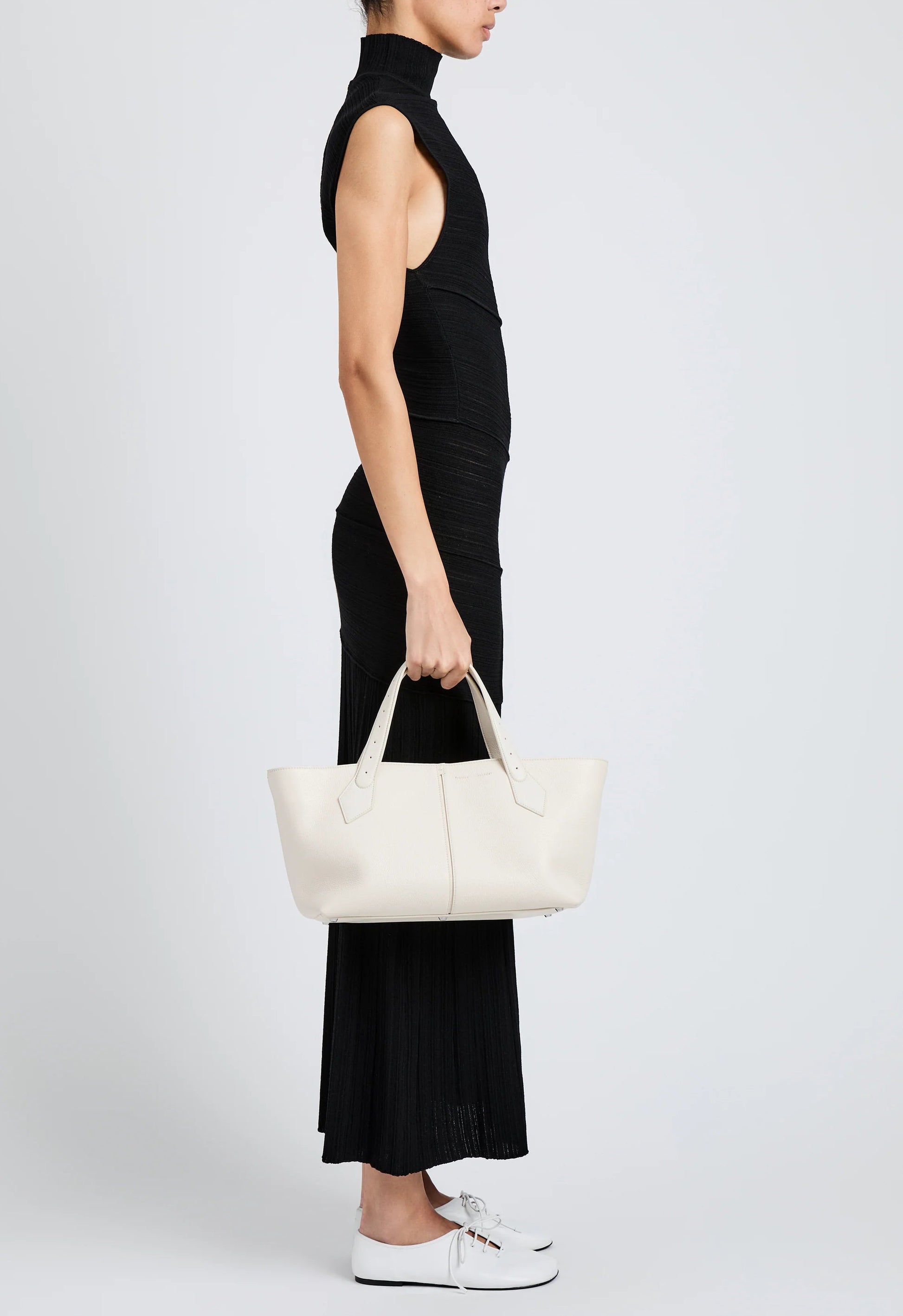 A person in a sleeveless black dress and white shoes stands in profile, holding the Proenza Schouler East West Chelsea Tote with two handles against a plain background.
