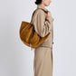 A person in a beige trench coat carries the Proenza Schouler Large Ruched Soft Suede Braided Tote, photographed from the side against a plain background.