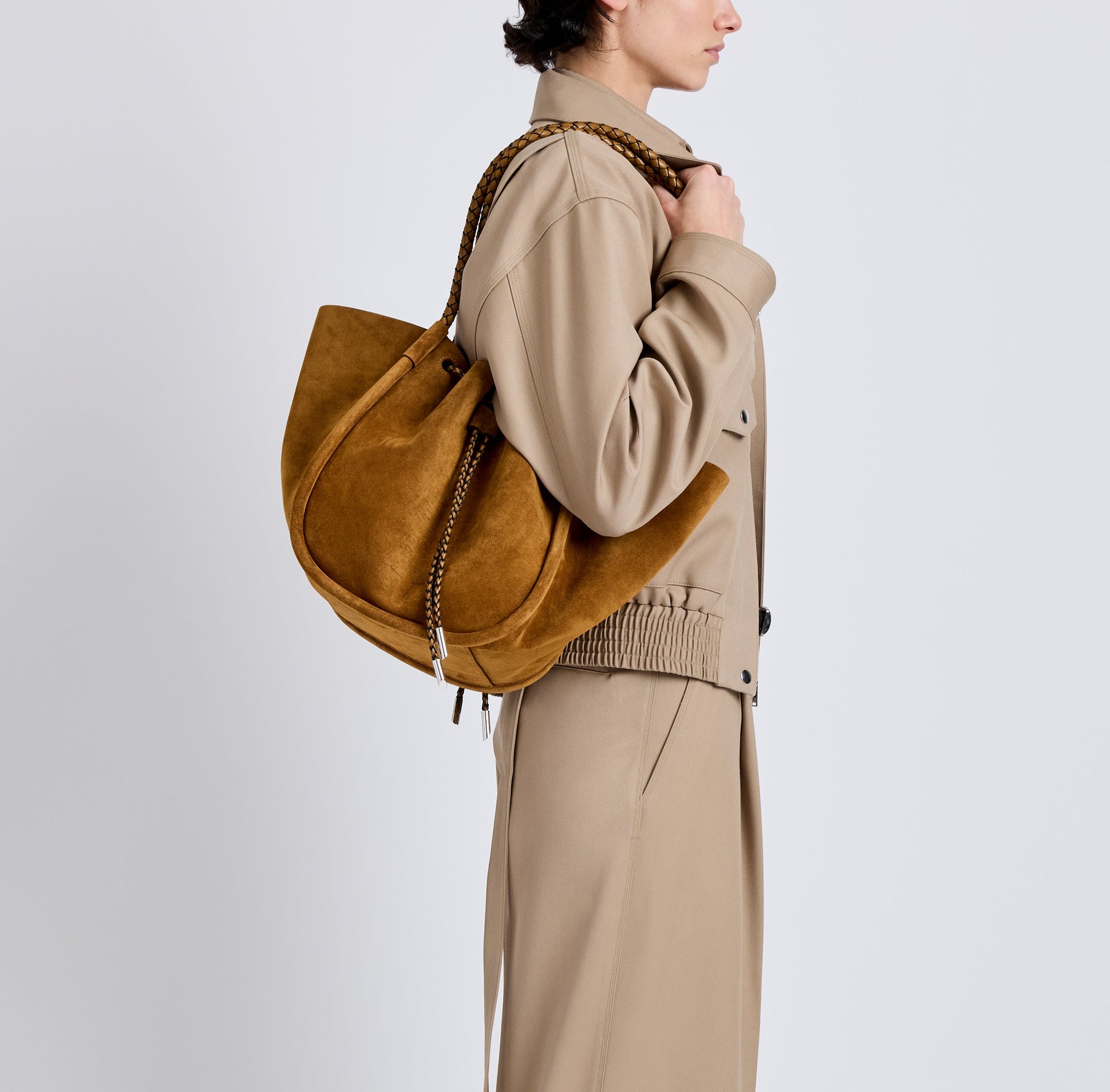 A person in a beige trench coat carries the Proenza Schouler Large Ruched Soft Suede Braided Tote, photographed from the side against a plain background.