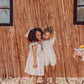 Two children wearing matching Louise Misha's Girls' Martine Dresses with embroidered floral details stand closely together, smiling, in front of a sunlit wall casting striped shadows.