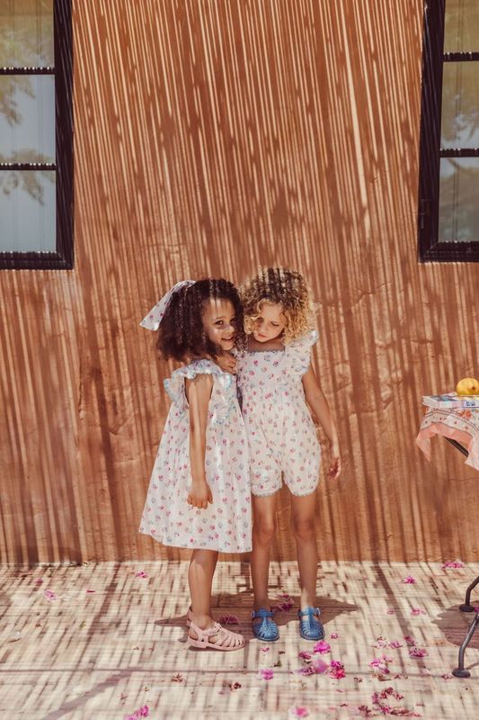 Two children wearing matching Louise Misha's Girls' Martine Dresses with embroidered floral details stand closely together, smiling, in front of a sunlit wall casting striped shadows.