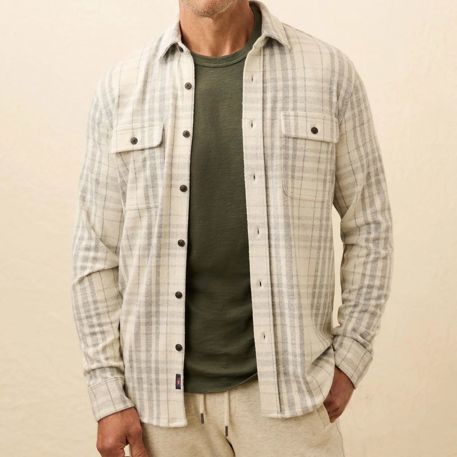 A man wears a Faherty Legend™ Sweater Shirt—a lightweight plaid button-up—over an olive green tee and beige pants, standing against a plain background.