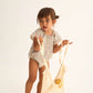 A young child wears the Cozmo Baby Girls' Holly Floral Bubble and holds a mesh bag with yellow objects, set against a plain light background.