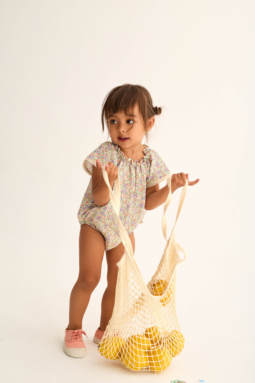 A young child wears the Cozmo Baby Girls' Holly Floral Bubble and holds a mesh bag with yellow objects, set against a plain light background.