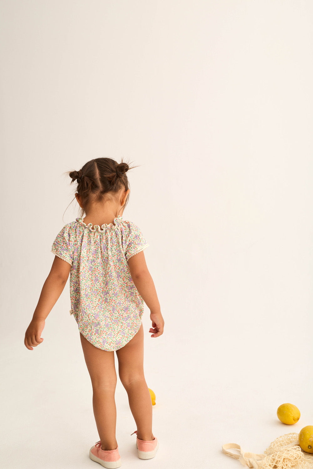 A young child with pigtails stands facing away, wearing the Cozmo Baby Girls' Holly Floral Bubble and pink shoes. Lemons and a net bag are on the white floor beside her.