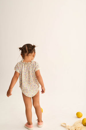 A young child with pigtails stands facing away, wearing the Cozmo Baby Girls' Holly Floral Bubble and pink shoes. Lemons and a net bag are on the white floor beside her.