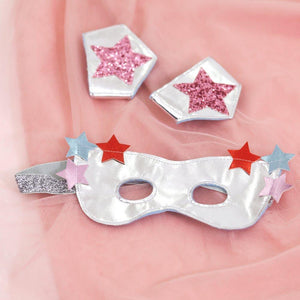 A shiny leatherette mask decorated with red and pink stars, placed on a pink fabric surface, accompanied by two smaller star-adorned accessories from the Meri Meri Superhero Dress Up Kit by Meri Meri.