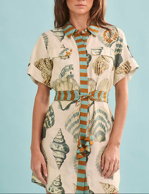 A woman models the Nicole Dress by Hunter Bell NYC—a short-sleeve mini dress with a seashell pattern and vertical orange and teal striped accents—against a light blue background.