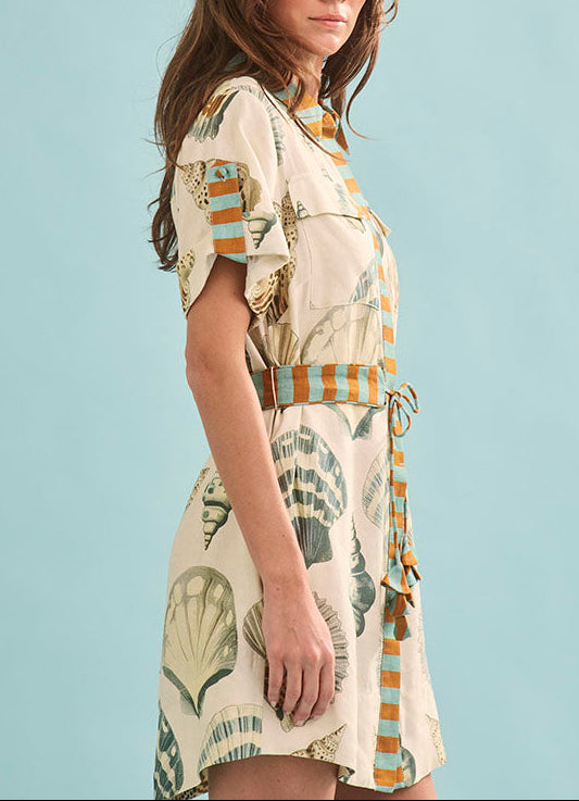 A woman stands against a plain light blue background wearing the Nicole Dress by Hunter Bell NYC, a short-sleeved, belted shirt dress featuring seashell and striped patterns.