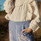 A child wearing a cream-colored, long-sleeved blouse with ruffles and light blue Louise Misha Girls’ Flofi Pants featuring delicate floral embroidery, standing outdoors.