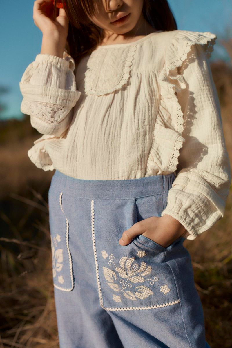 A child wearing a cream-colored, long-sleeved blouse with ruffles and light blue Louise Misha Girls’ Flofi Pants featuring delicate floral embroidery, standing outdoors.
