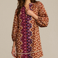A woman wears the Hailey Dress by Figue, a long-sleeve cotton poplin dress featuring geometric designs in brown, orange, and purple with a subtle band collar.