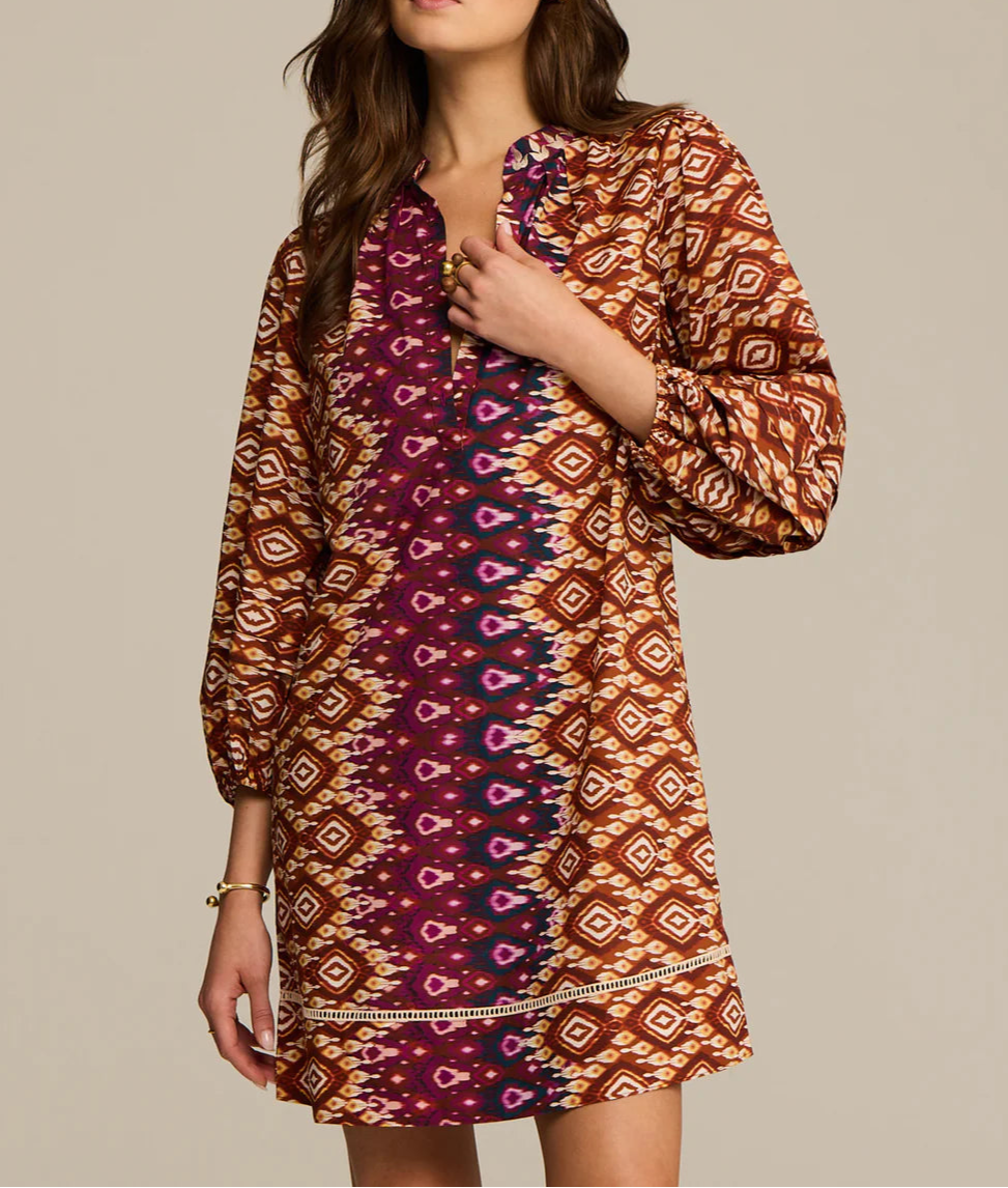 A woman wears the Hailey Dress by Figue, a long-sleeve cotton poplin dress featuring geometric designs in brown, orange, and purple with a subtle band collar.