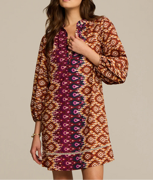 A woman wears the Hailey Dress by Figue, a long-sleeve cotton poplin dress featuring geometric designs in brown, orange, and purple with a subtle band collar.