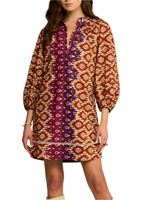 A woman wears the Hailey Dress by Figue—a knee-length cotton poplin dress with geometric and ikat patterns in brown, orange, and magenta, featuring balloon sleeves, a band collar, and a split neckline.