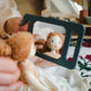 In a delightful hair salon setup, a person holds the *Tender Leaf Hair Salon* wooden doll from *Tender Leaf Toys* in front of a handheld mirror, its face reflected with various styling accessories scattered around. The scene is set on a quilt adorned with red and white flowers, enhancing the charm of this playful arrangement.