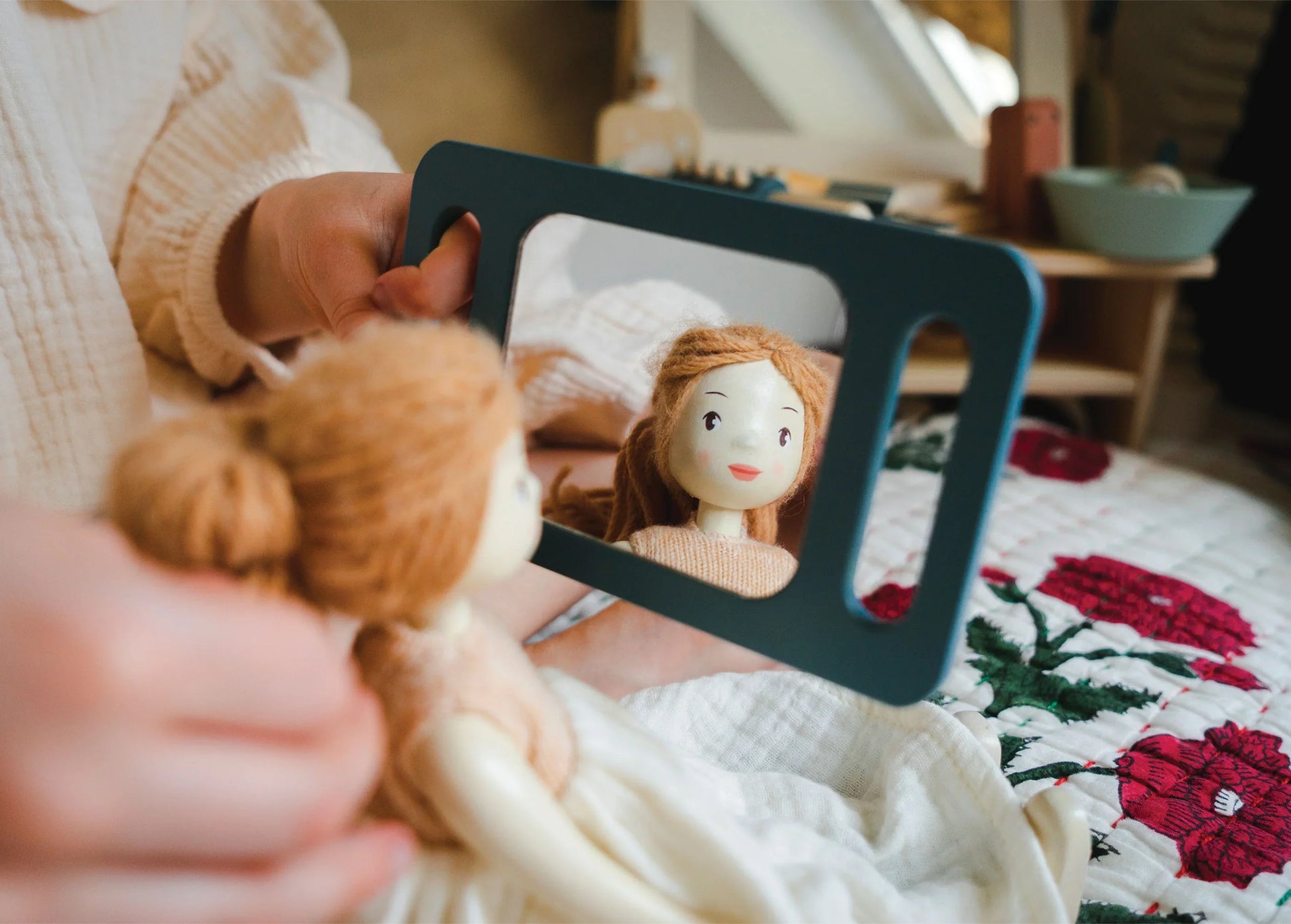 In a delightful hair salon setup, a person holds the *Tender Leaf Hair Salon* wooden doll from *Tender Leaf Toys* in front of a handheld mirror, its face reflected with various styling accessories scattered around. The scene is set on a quilt adorned with red and white flowers, enhancing the charm of this playful arrangement.